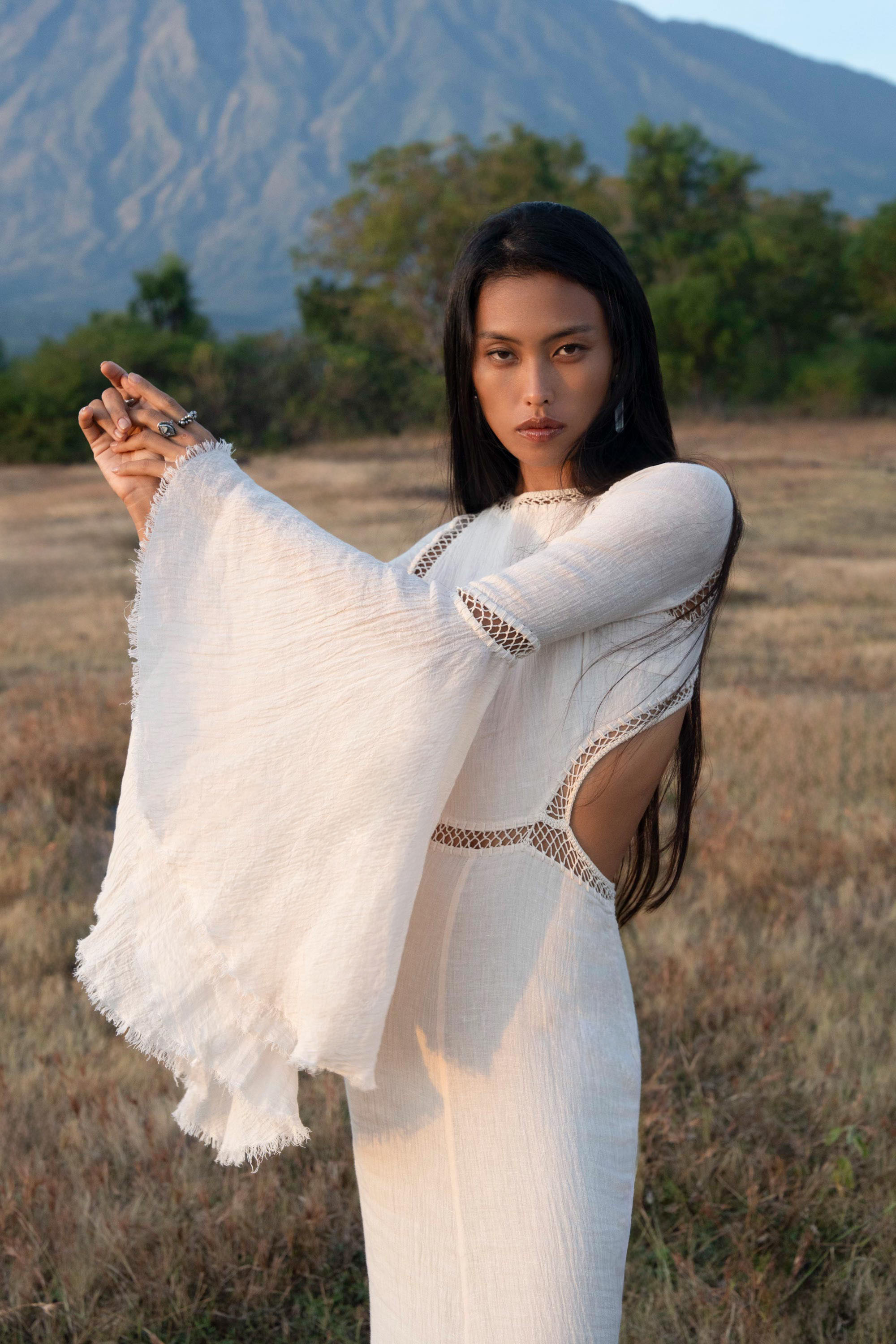 A woman with long dark hair stands in a field, wearing the Mandara Dress in ivory by AYA Sacred Wear—an organic boho wedding dress with open sleeves and side cut-outs—arms raised, mountains and greenery behind her under a clear sky.