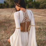 A woman with long dark hair stands in a field, wearing the Mandara Dress in Ivory by AYA Sacred Wear—an organic boho wedding dress with wide bell sleeves and hand-stitched embroidery. Green trees and a mountain are visible under a clear blue sky.
