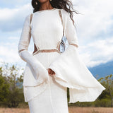 A woman stands in a field wearing the Mandara Dress in Ivory by AYA Sacred Wear—an organic boho wedding dress with linen crinkle fabric, bell sleeves, cut-outs, and hand-stitched embroidery. Her dark wavy hair is loose as she poses amid blurred greenery.