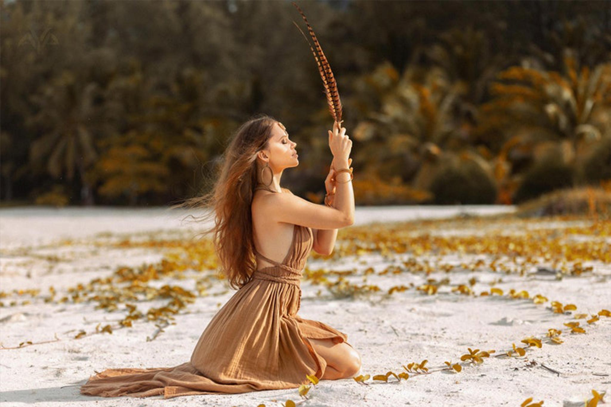 A woman in an Ochre Greek Goddess Dress by AYA Sacred Wear kneels on a sandy beach, holding a feather upright in her hands. She is surrounded by green vegetation, with lush palm trees in the background, creating a scene that conveys a sense of peace and connection with nature.