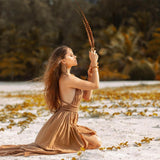 A woman in an Ochre Greek Goddess Dress by AYA Sacred Wear kneels on a sandy beach, holding a feather upright in her hands. She is surrounded by green vegetation, with lush palm trees in the background, creating a scene that conveys a sense of peace and connection with nature.