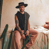 A bearded man sits on a cloth-covered surface surrounded by plants, wearing a wide-brimmed hat and sleeveless black top. He dons Ochre Orange Long Cotton Pants for Men from AYA Sacred Wear, with a patterned scarf draped over one shoulder, in a setting with warm, rustic ambiance and earthy tones.