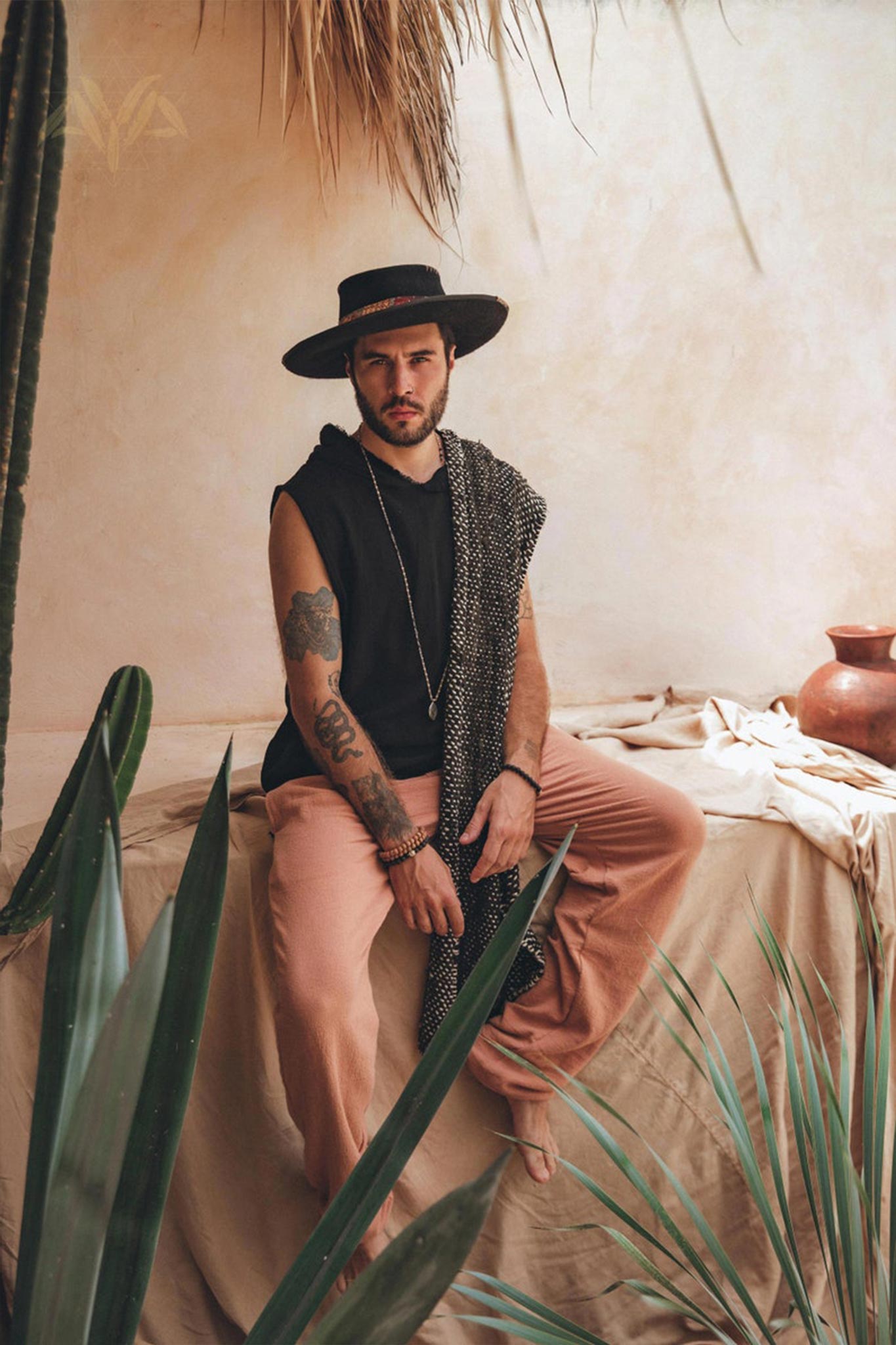 A bearded man sits on a cloth-covered surface surrounded by plants, wearing a wide-brimmed hat and sleeveless black top. He dons Ochre Orange Long Cotton Pants for Men from AYA Sacred Wear, with a patterned scarf draped over one shoulder, in a setting with warm, rustic ambiance and earthy tones.