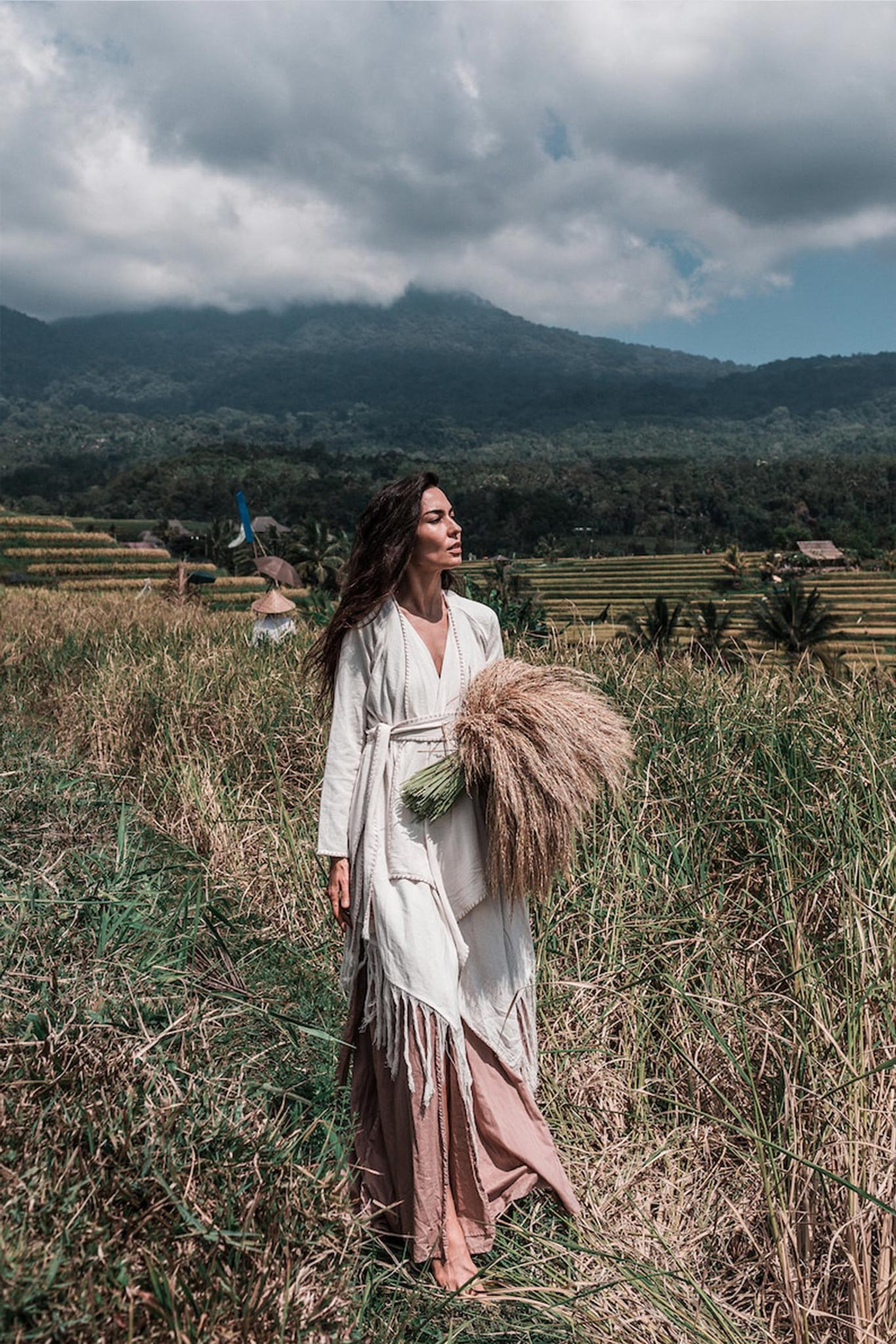 A woman in an Off-White Bohemian Raw Cotton Coat Cape Wrap from AYA Sacred Wear strides through a grassy field, clutching a large bundle of dried grass. Layered rice terraces and a mountain beneath a cloudy sky form the backdrop.