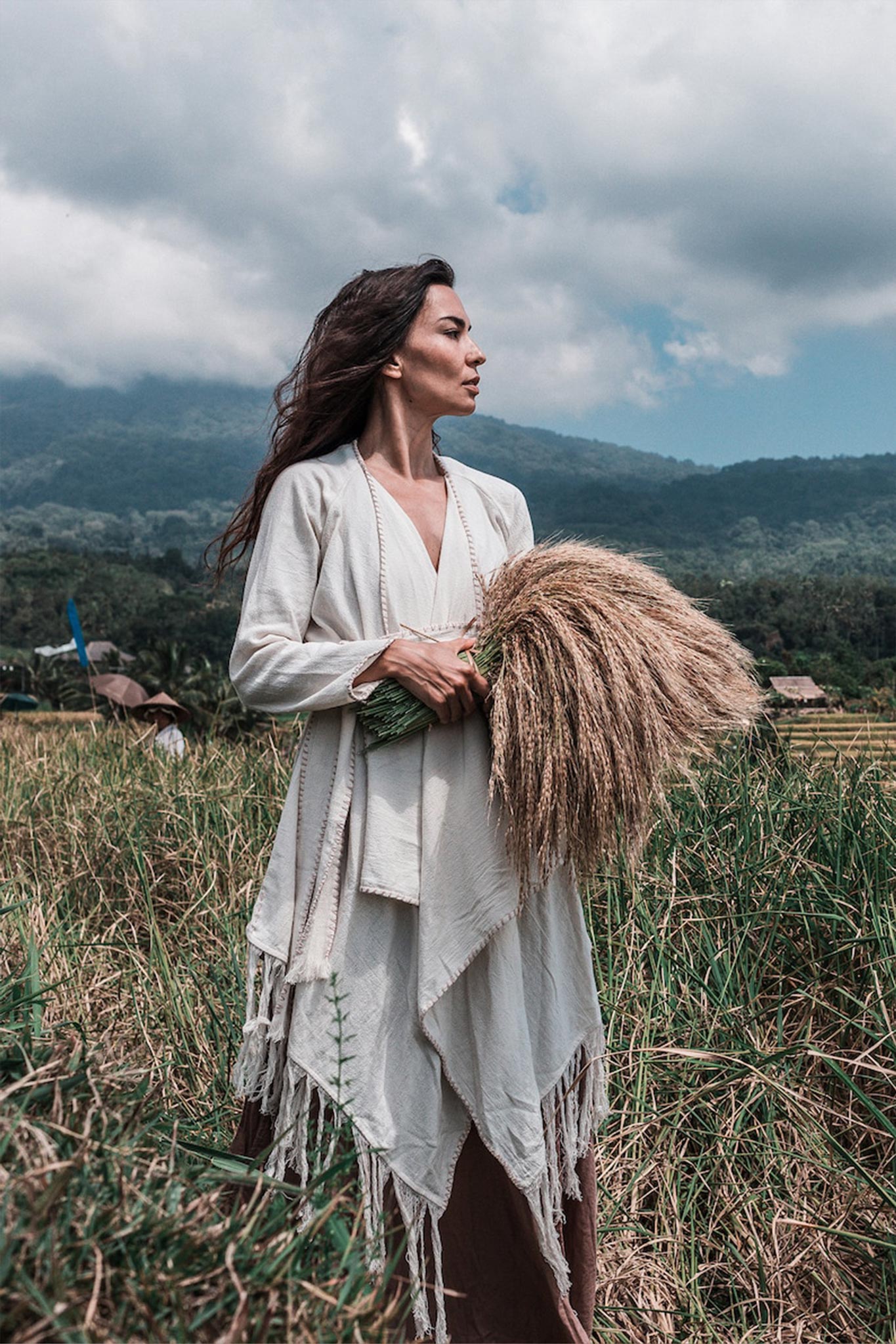 A woman stands in a field, holding a bundle of wheat. She wears an Off-White Bohemian Raw Cotton Coat Cape Wrap by AYA Sacred Wear, featuring exquisite hand stitch embroidery, and gazes into the distance. The background highlights lush greenery and a cloudy sky over distant hills.