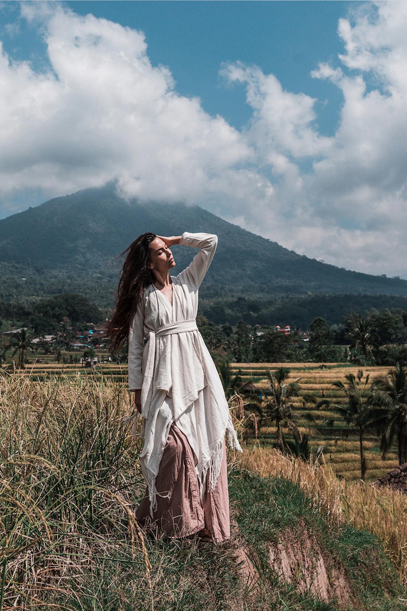 A woman in an Off-White Bohemian Raw Cotton Coat Cape Wrap by AYA Sacred Wear stands on a grassy ridge, gazing upward with her hand shielding her eyes. A lush mountain and fields extend into the background under a partly cloudy sky, creating the perfect setting for her cape to catch the gentle breeze.