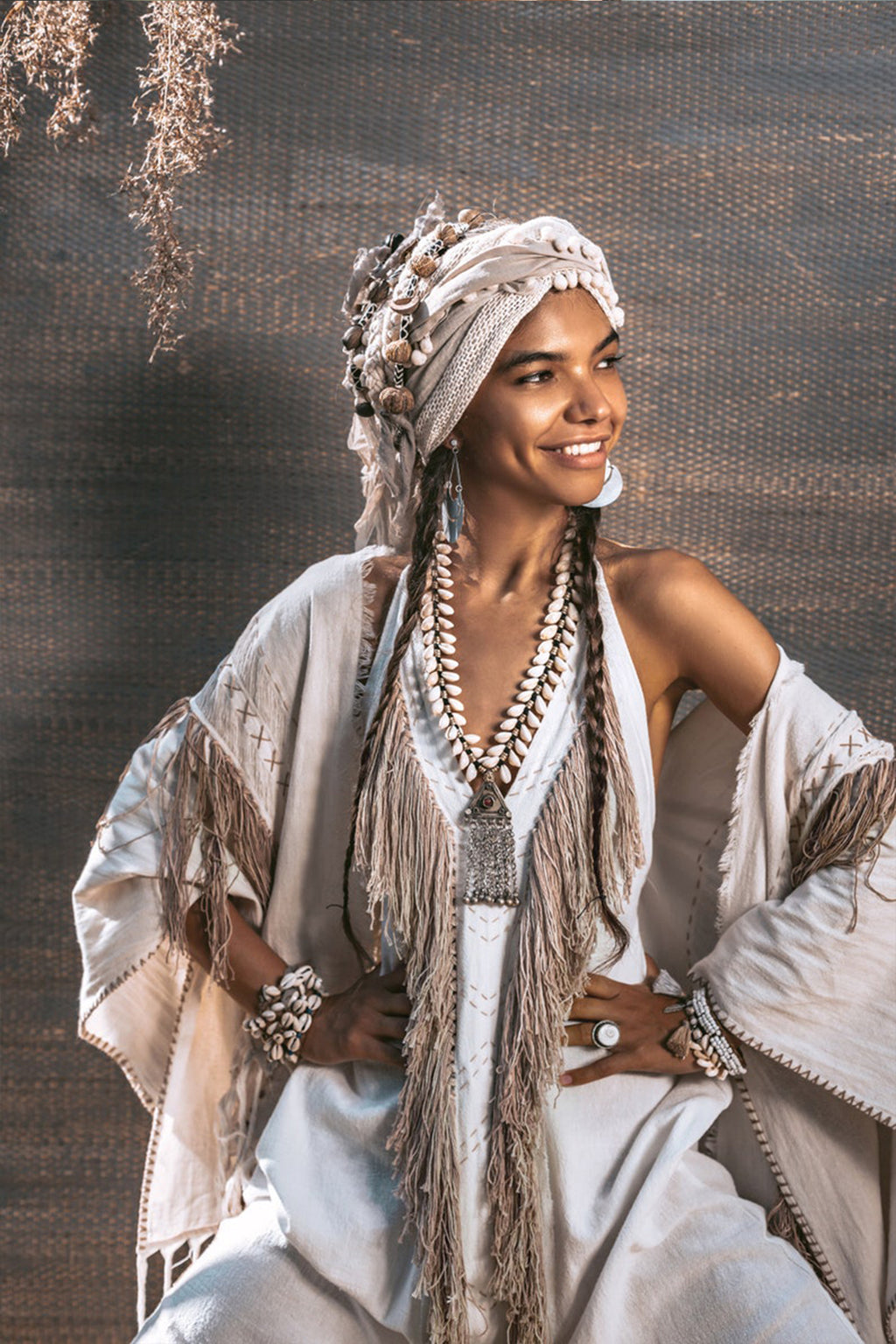 A person wearing an off-white boho tribal bohemian dress from AYA Sacred Wear, complete with a fringed, light-colored outfit and a headwrap embellished with shells, smiles while posing. The handmade bridesmaid ensemble is complemented by layered necklaces and bracelets as they stand with their hands on their hips against a textured backdrop.