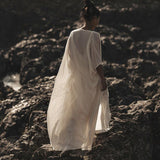 An individual draped in an Off-White Cotton Boho Kimono Cover-up by AYA Sacred Wear stands gracefully on rocky terrain. This exquisite piece, functioning as a bridal cape or poncho robe, enhances the serene and mysterious atmosphere with its sustainable fashion flair against a backdrop of rugged, dark rocks.