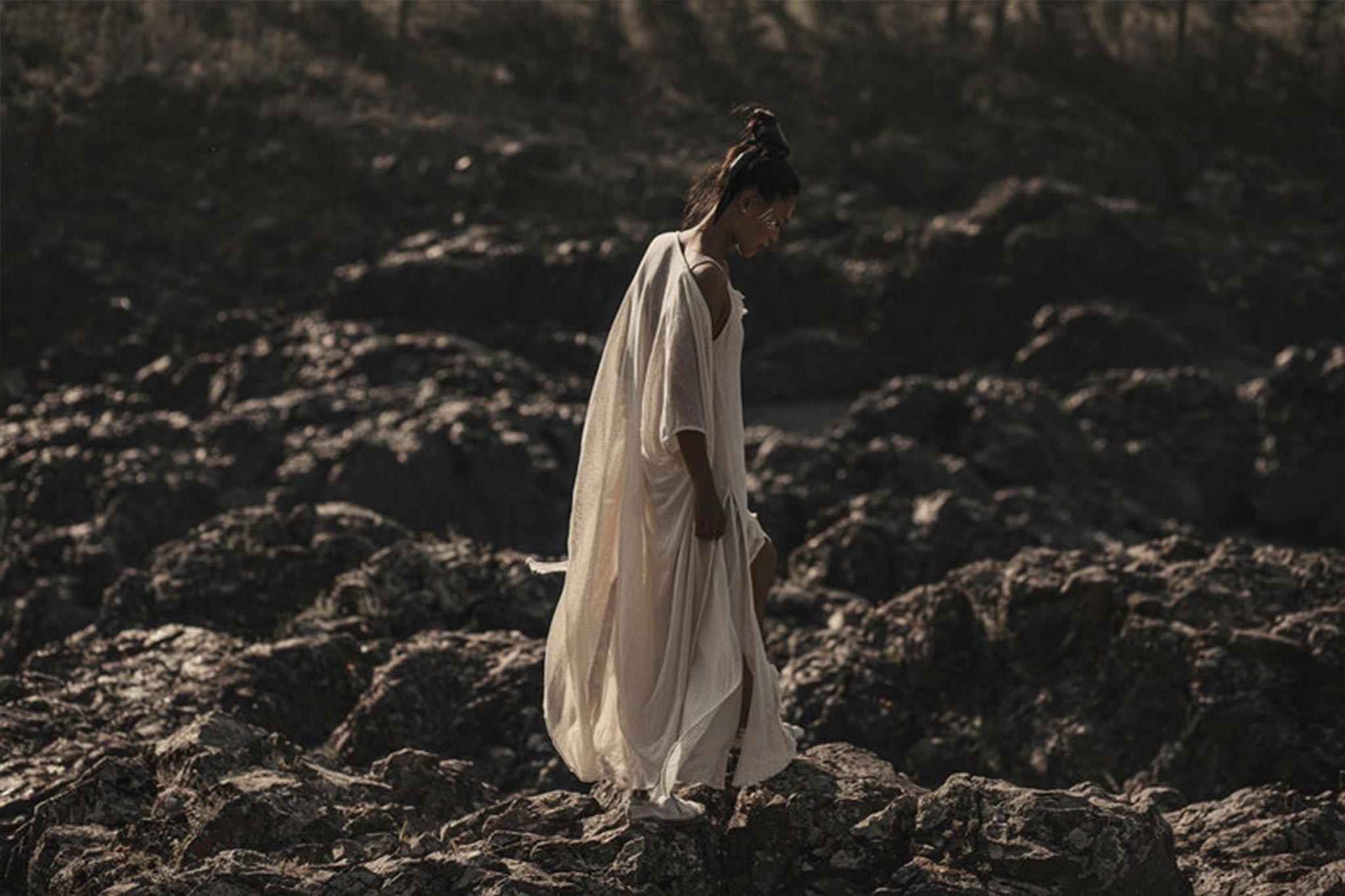 A person wearing the Off-White Cotton Boho Kimono Cover-up by AYA Sacred Wear walks over rugged, rocky terrain. The muted colors and natural landscape create a serene and contemplative atmosphere, embodying the essence of sustainable fashion.