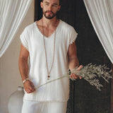 A person with a beard stands in a relaxed pose, donning the Off-White Handmade Knots Decorated T-Shirt for Men from AYA Sacred Wear. They hold dried flowers in one hand, surrounded by draped curtains and a large vase.
