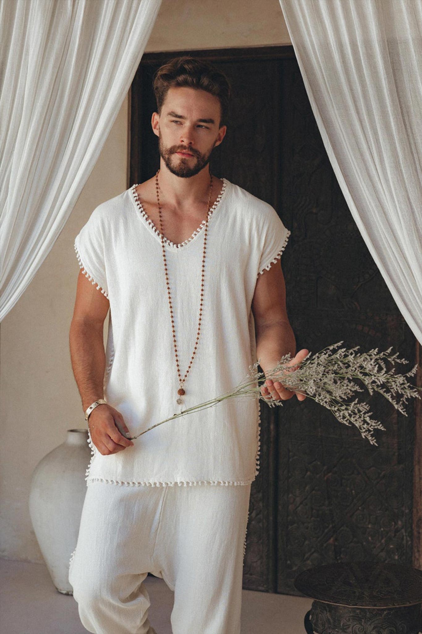 A person with a beard stands in a relaxed pose, donning the Off-White Handmade Knots Decorated T-Shirt for Men from AYA Sacred Wear. They hold dried flowers in one hand, surrounded by draped curtains and a large vase.