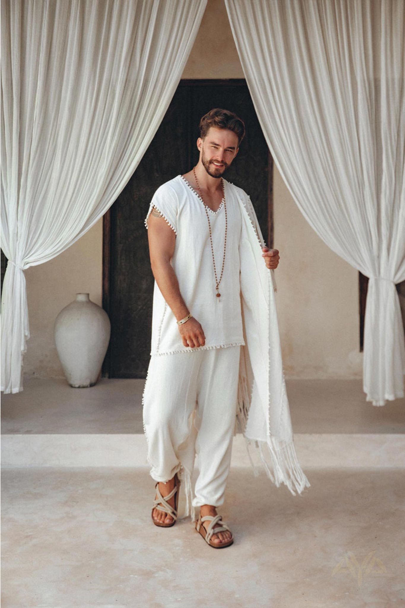 A person stands on a light-colored floor, wearing an Off-White Handmade Knots Decorated T-Shirt by AYA Sacred Wear and botanically dyed pants, accessorized with a necklace and sandals. They are positioned in front of open white curtains, revealing a dark doorway and a large ceramic vase.