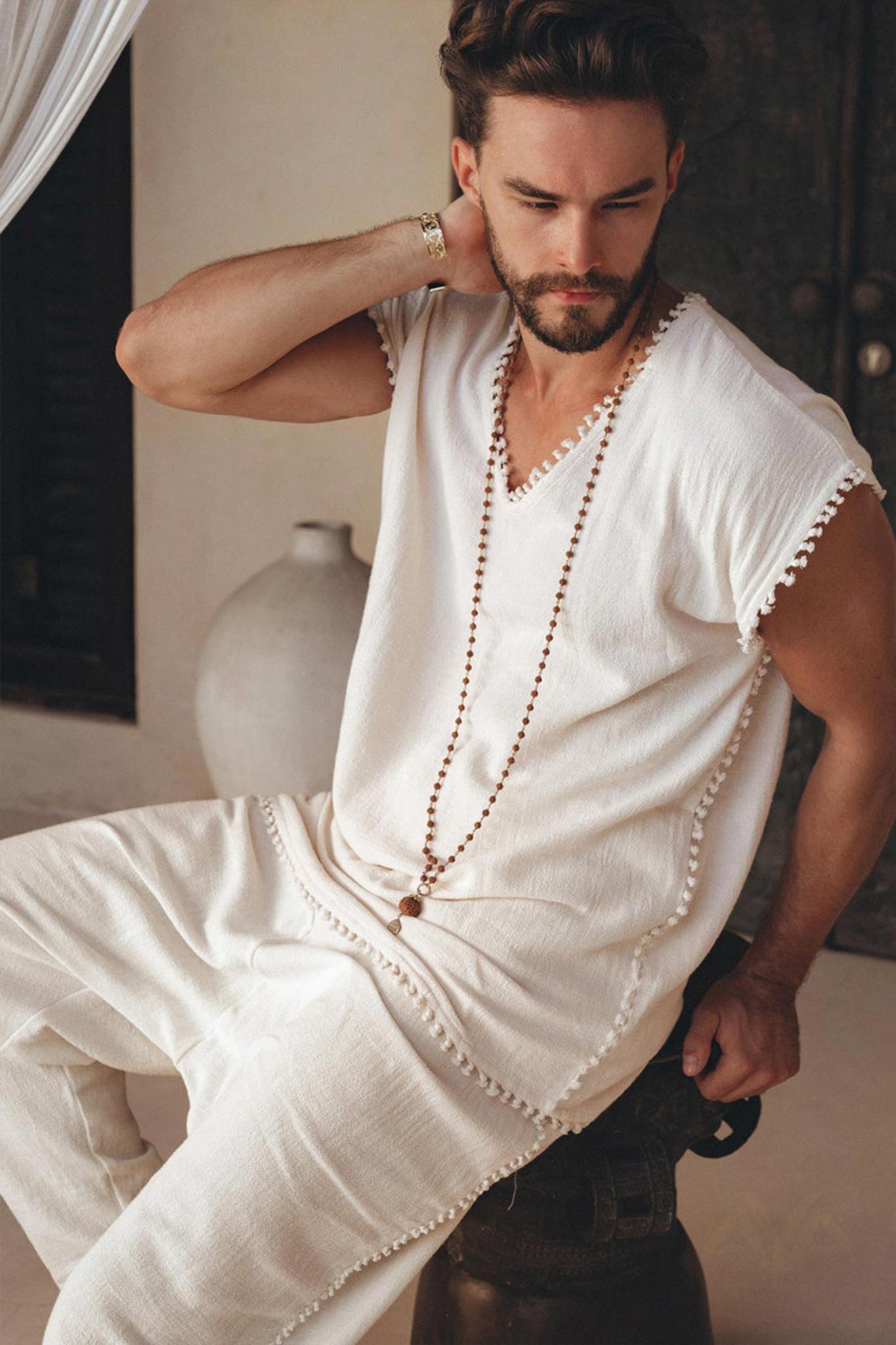 A man with a beard sits on a stool wearing an off-white handmade knots-decorated T-shirt from AYA Sacred Wear. Crafted from handwoven organic cotton, this boho-style shirt perfectly complements his long beaded necklace. He gently touches his neck against a backdrop featuring a large ceramic pot.