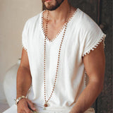 A bearded man wearing an Off-White Handmade Knots Decorated T-Shirt for Men from AYA Sacred Wear sits while holding a small branch with delicate leaves. The background features a textured surface with a muted color palette, enhancing the boho aesthetic of his long necklace and natural, botanically dyed attire.