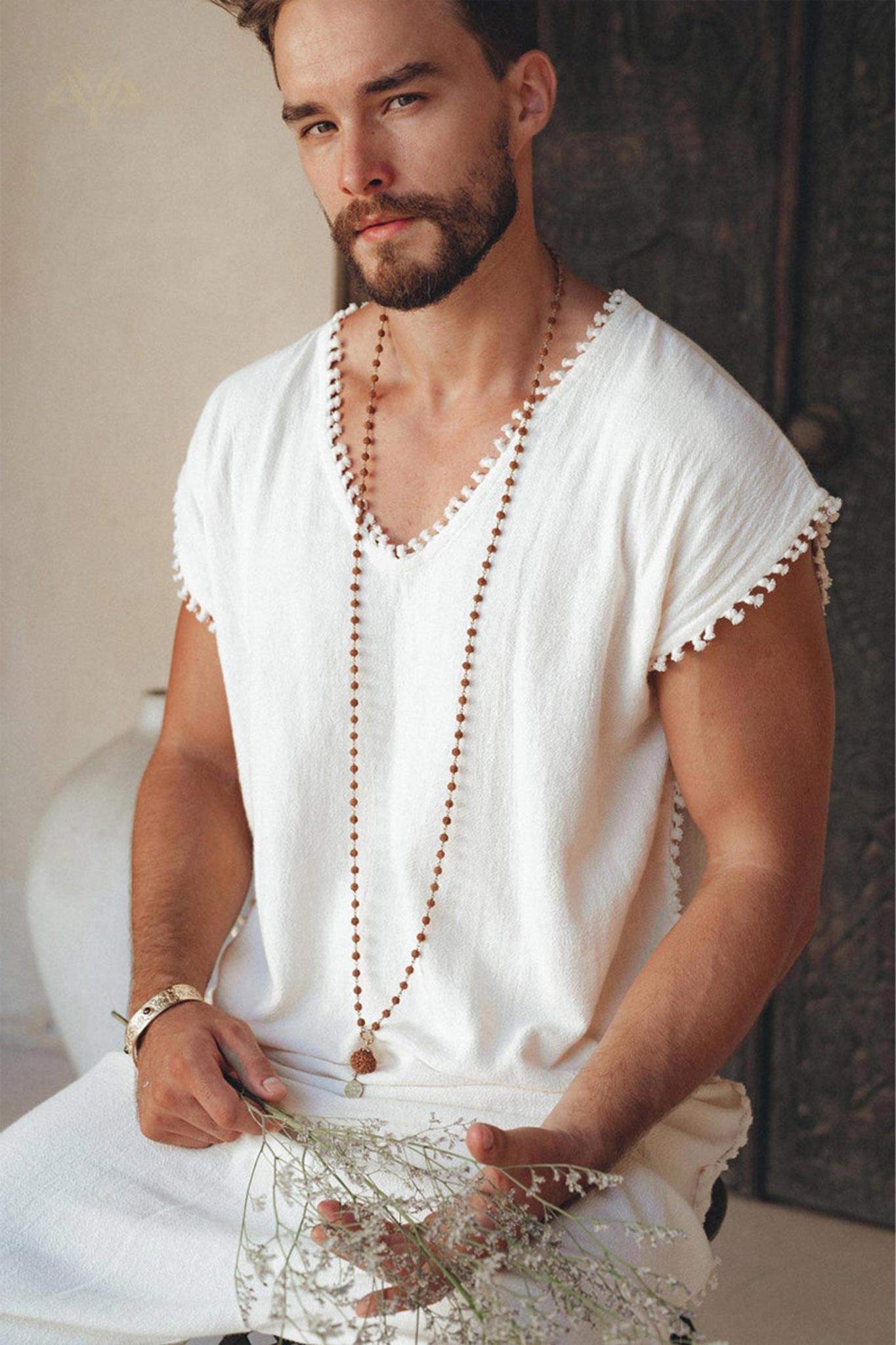 A bearded man wearing an Off-White Handmade Knots Decorated T-Shirt for Men from AYA Sacred Wear sits while holding a small branch with delicate leaves. The background features a textured surface with a muted color palette, enhancing the boho aesthetic of his long necklace and natural, botanically dyed attire.