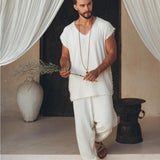 A man with a beard stands in a relaxed pose, dressed in an off-white handmade knots decorated T-shirt by AYA Sacred Wear and loose-fitting pants. He holds a sprig of dried flowers, with sheer white curtains in the background, and a large vase and metallic stool nearby.