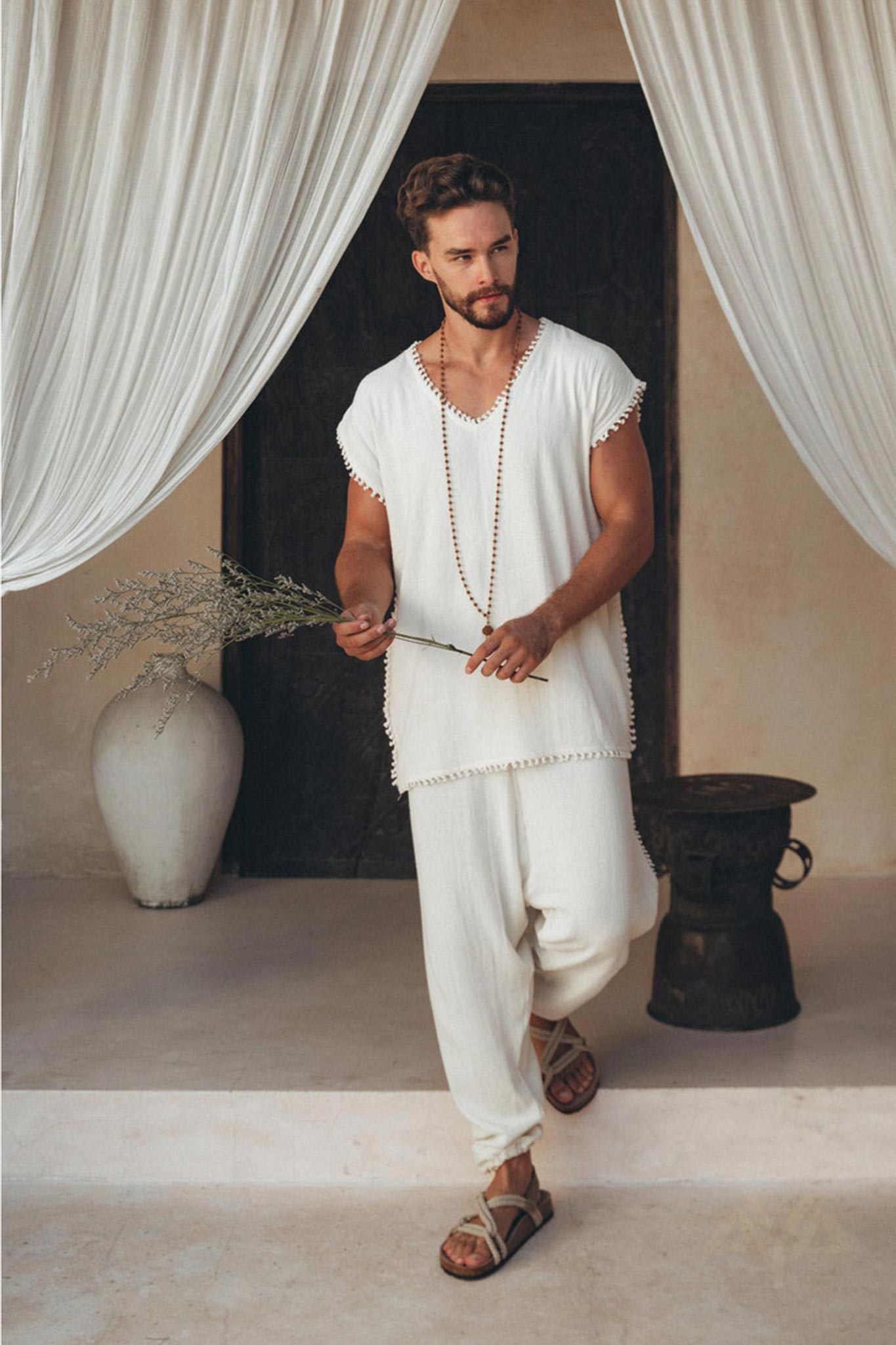 A man with a beard stands in a relaxed pose, dressed in an off-white handmade knots decorated T-shirt by AYA Sacred Wear and loose-fitting pants. He holds a sprig of dried flowers, with sheer white curtains in the background, and a large vase and metallic stool nearby.