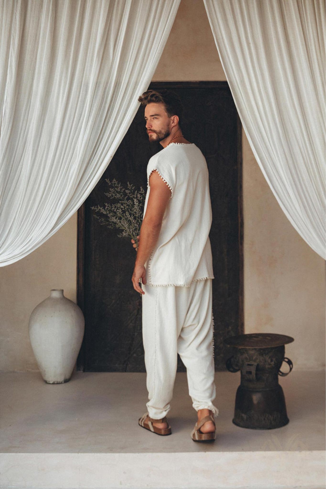 In an elegant room, a bearded man holds a bouquet of botanically dyed flowers while dressed in an Off-White Handmade Knots Decorated T-Shirt from AYA Sacred Wear, along with loose-fitting pants made from handwoven organic cotton and sandals. The area is adorned with white drapes framing a dark wooden door, and a large white vase rests on the floor.