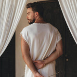 A man wearing an Off-White Handmade Knots Decorated T-Shirt by AYA Sacred Wear stands with his back to the camera, holding dried flowers behind him. He is positioned between two white curtains and in front of a dark, textured door.