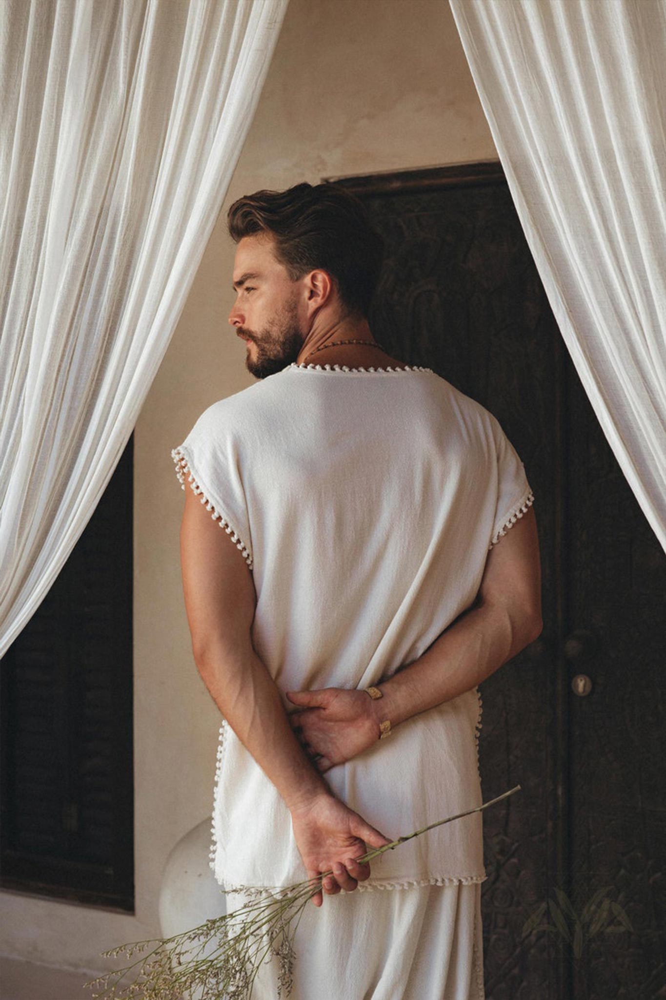 A man wearing an Off-White Handmade Knots Decorated T-Shirt by AYA Sacred Wear stands with his back to the camera, holding dried flowers behind him. He is positioned between two white curtains and in front of a dark, textured door.