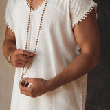 A man wearing an AYA Sacred Wear Off-White Handmade Knots Decorated T-Shirt holds a long beaded necklace. The image highlights the upper body, emphasizing the simple and elegant apparel and jewelry in a tranquil setting.