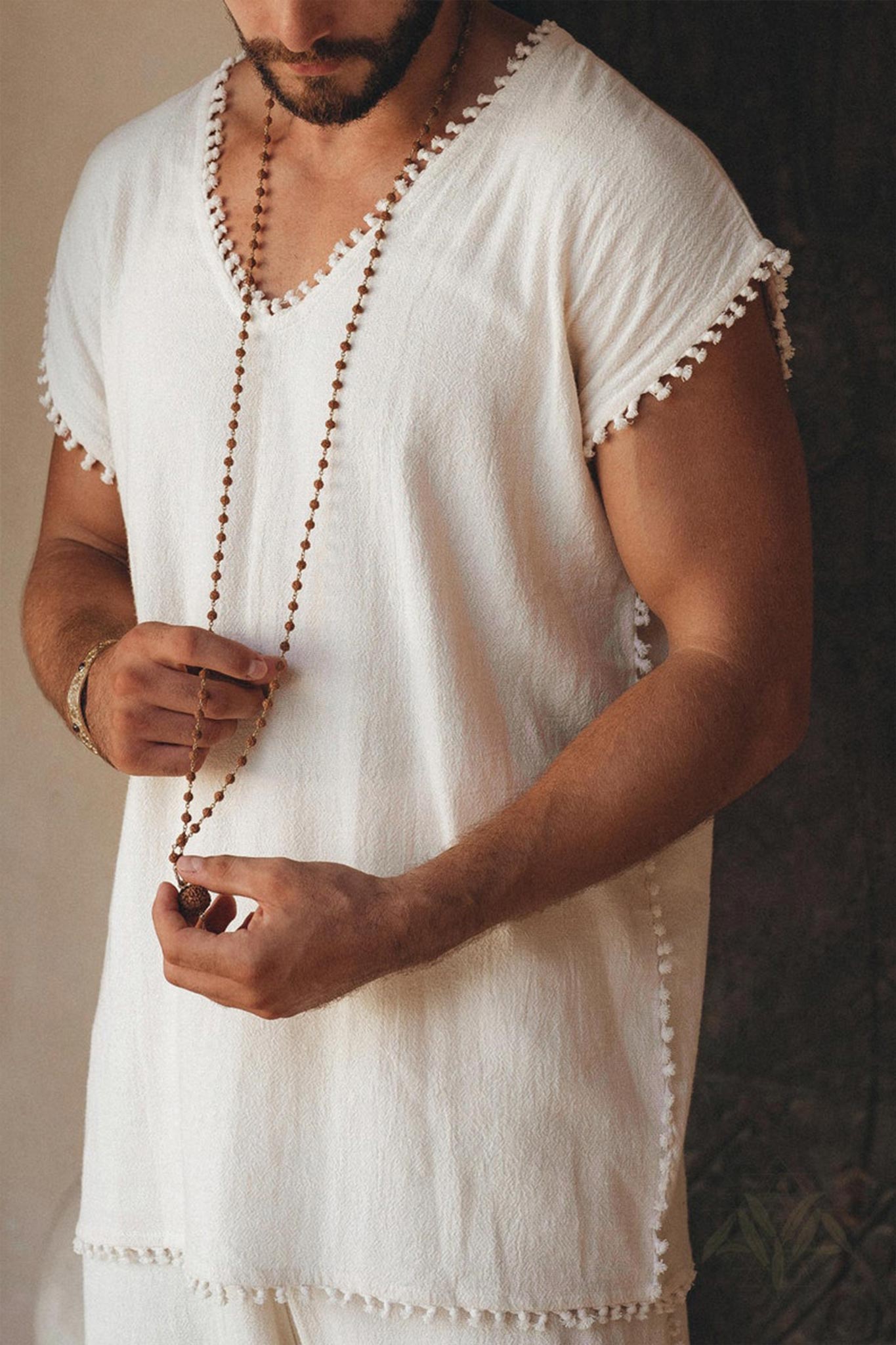 A man wearing an AYA Sacred Wear Off-White Handmade Knots Decorated T-Shirt holds a long beaded necklace. The image highlights the upper body, emphasizing the simple and elegant apparel and jewelry in a tranquil setting.