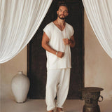 In a tranquil environment, an individual stands outfitted in the Off-White Handmade Knots Decorated T-Shirt for Men from AYA Sacred Wear, made of handwoven organic cotton with brown accents. Holding a string of beads and wearing sandals, they are surrounded by white drapes with a large vase and decorative stool nearby.