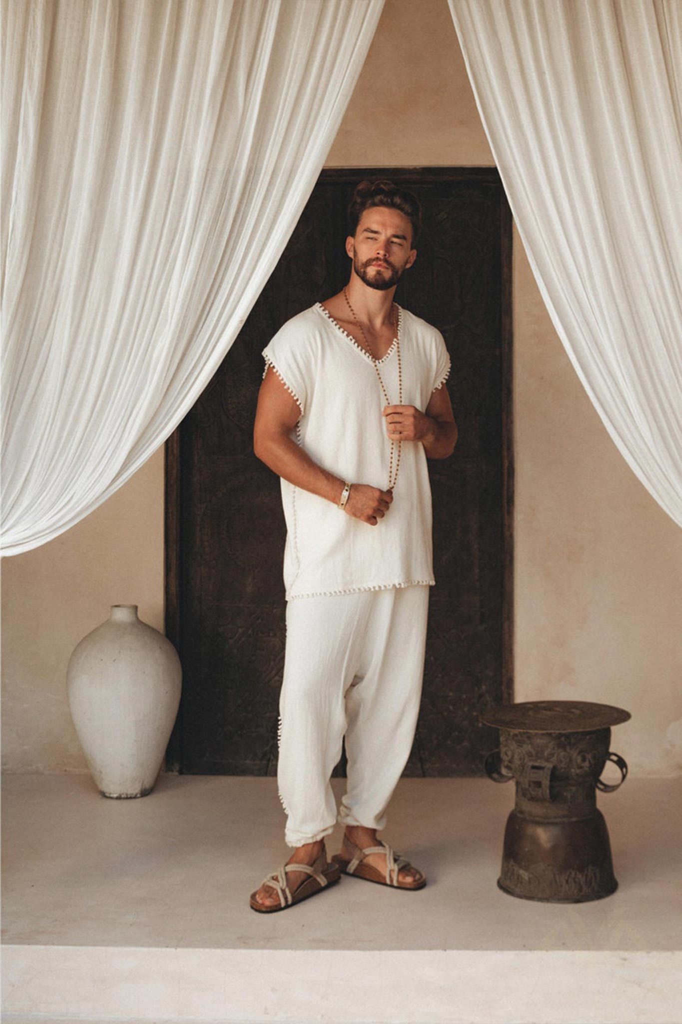 In a tranquil environment, an individual stands outfitted in the Off-White Handmade Knots Decorated T-Shirt for Men from AYA Sacred Wear, made of handwoven organic cotton with brown accents. Holding a string of beads and wearing sandals, they are surrounded by white drapes with a large vase and decorative stool nearby.