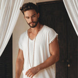 A person with a beard and short hair stands in front of a dark wooden door, wearing an Off-White Handmade Knots Decorated T-Shirt for Men by AYA Sacred Wear. A long chain necklace hangs down, while white curtains frame the scene.