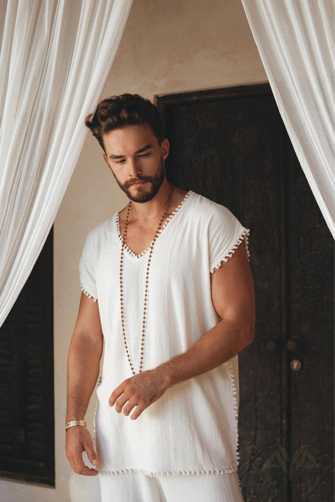 A person with a beard and short hair stands in front of a dark wooden door, wearing an Off-White Handmade Knots Decorated T-Shirt for Men by AYA Sacred Wear. A long chain necklace hangs down, while white curtains frame the scene.