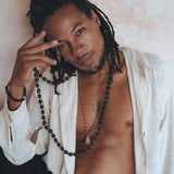 A person with long, braided hair poses against a textured wall, wearing the Off-White Handwoven Cotton Oversized Shirt for Men by AYA Sacred Wear. The shirt is slightly open to reveal a beaded necklace. They look directly at the camera while holding part of the necklace in their hand.