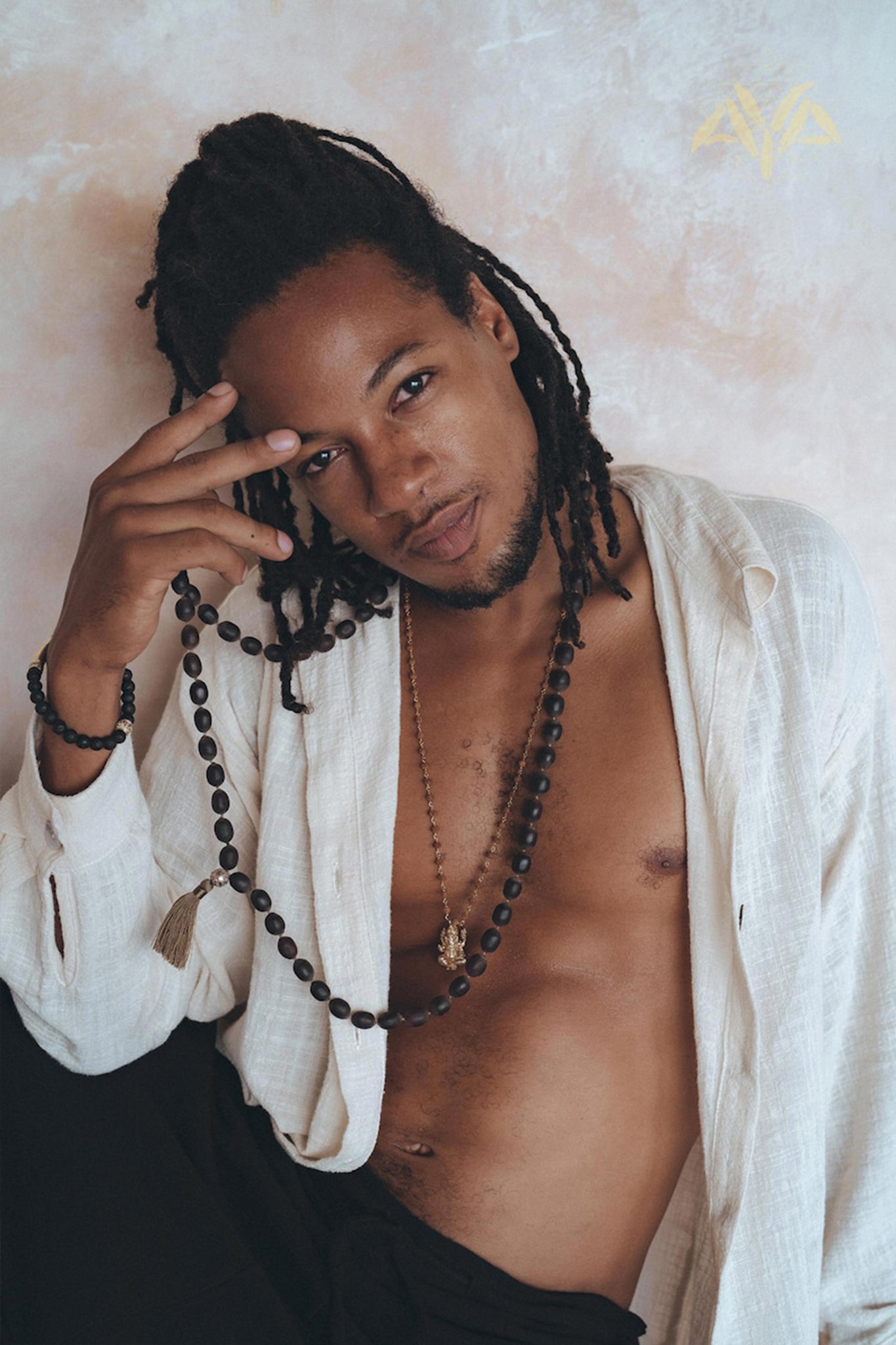 A person with long, braided hair poses against a textured wall, wearing the Off-White Handwoven Cotton Oversized Shirt for Men by AYA Sacred Wear. The shirt is slightly open to reveal a beaded necklace. They look directly at the camera while holding part of the necklace in their hand.