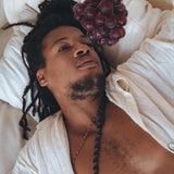 A person with long hair relaxes on a bed wearing an oversized, handwoven Off-White Cotton Shirt from AYA Sacred Wear. A beaded necklace adorns their neck as they casually hold a bunch of red grapes above them, capturing the essence of natural elegance.