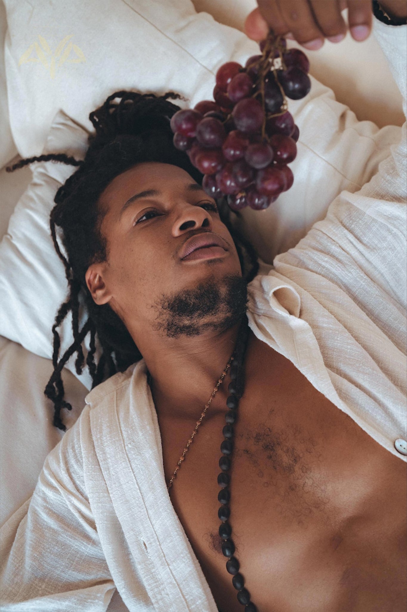 A person with long hair relaxes on a bed wearing an oversized, handwoven Off-White Cotton Shirt from AYA Sacred Wear. A beaded necklace adorns their neck as they casually hold a bunch of red grapes above them, capturing the essence of natural elegance.