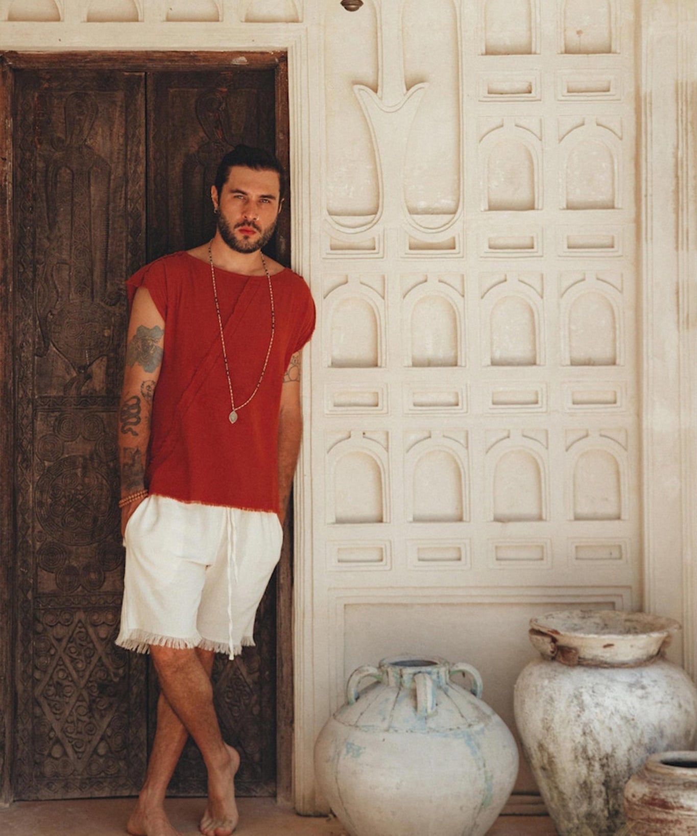 A tattooed man stands barefoot against a carved wooden door, wearing an AYA Sacred Wear red shirt and Off-White Handwoven Cotton Shorts for Men. In a rustic setting adorned with ornate wall designs and large ceramic pots, he radiates an earthy charm in his botanically dyed outfit.