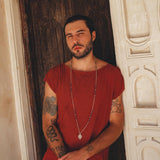 A man with short hair and a beard leans against a carved wooden door wearing a red shirt, long necklace, and AYA Sacred Wear's Off-White Handwoven Cotton Shorts for Men. His arms feature several tattoos, while the ornate white architectural details in the background enhance his earthy charm.