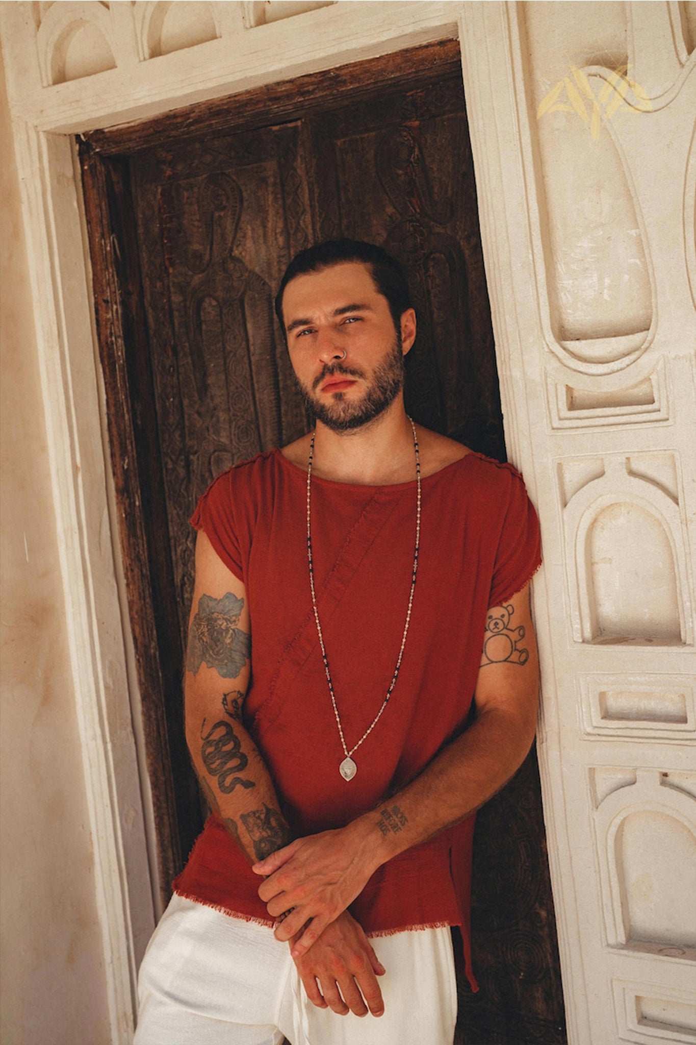 A man with short hair and a beard leans against a carved wooden door wearing a red shirt, long necklace, and AYA Sacred Wear's Off-White Handwoven Cotton Shorts for Men. His arms feature several tattoos, while the ornate white architectural details in the background enhance his earthy charm.