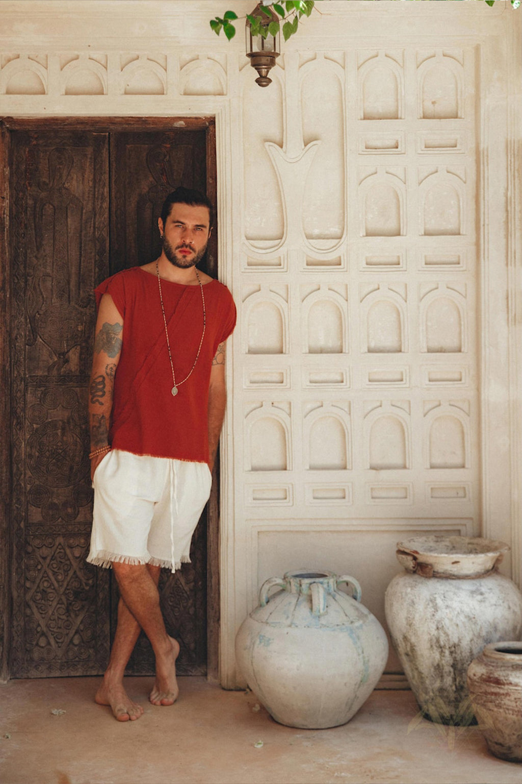 A tattooed man stands barefoot against a carved wooden door, wearing an AYA Sacred Wear red shirt and Off-White Handwoven Cotton Shorts for Men. In a rustic setting adorned with ornate wall designs and large ceramic pots, he radiates an earthy charm in his botanically dyed outfit.