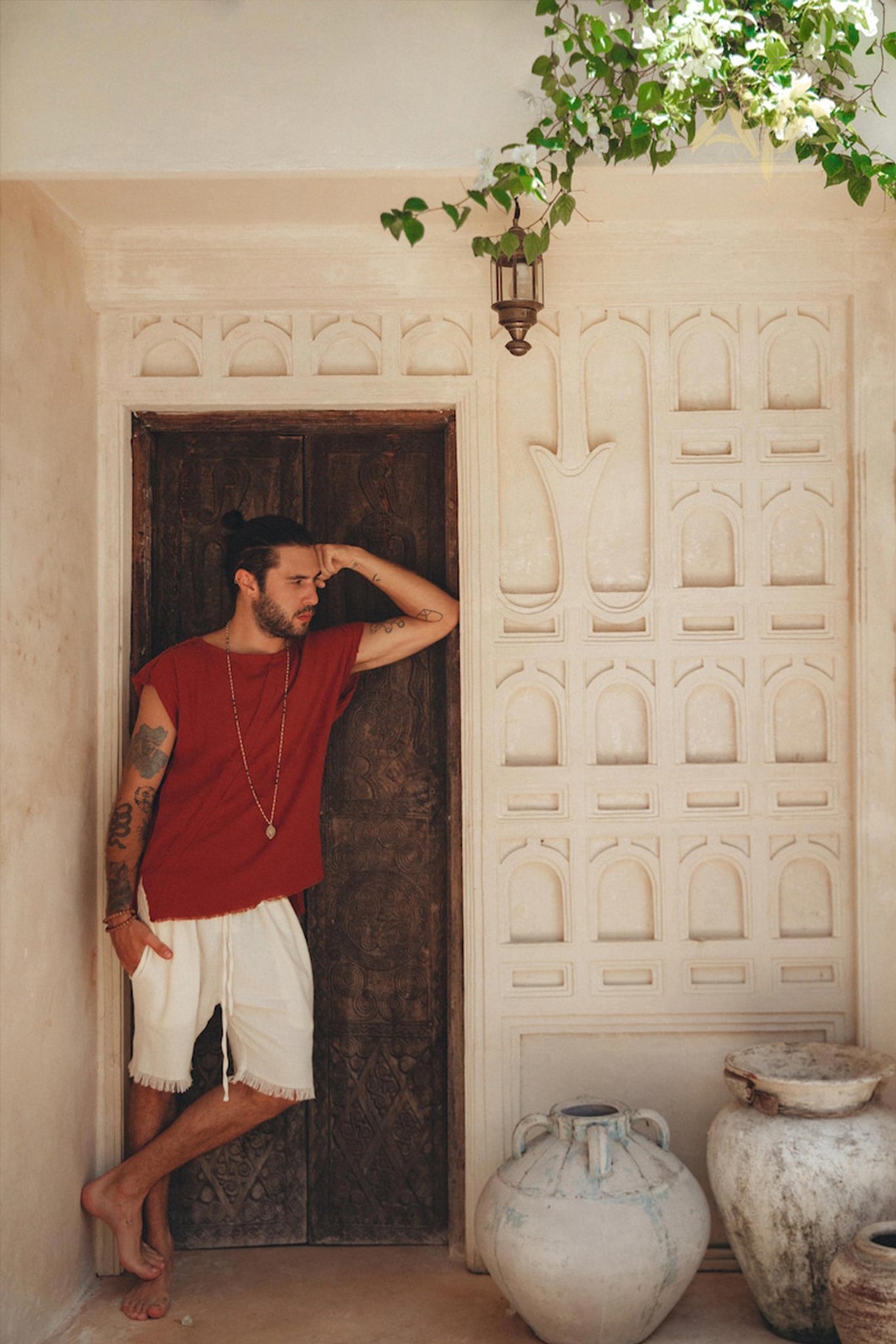 A man with tattoos leans against a carved wooden door, wearing a red shirt and Off-White Handwoven Cotton Shorts for Men by AYA Sacred Wear. Potted plants and ornate vases are placed nearby. A lantern hangs above, and a flowering vine adds greenery to the scene.