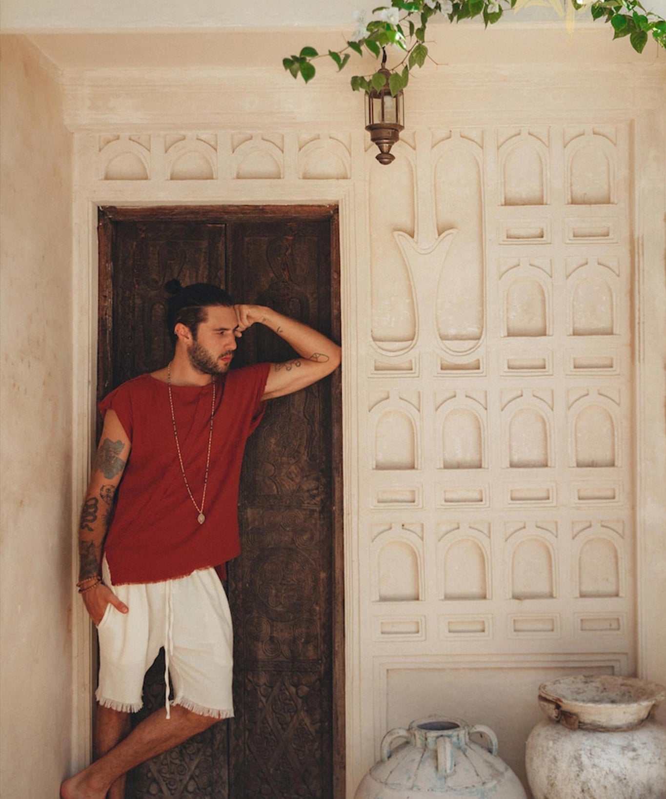 A man with tattoos leans against a carved wooden door, wearing a red shirt and Off-White Handwoven Cotton Shorts for Men by AYA Sacred Wear. Potted plants and ornate vases are placed nearby. A lantern hangs above, and a flowering vine adds greenery to the scene.