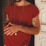A bearded man with tattoos stands against a textured wooden wall, wearing an outfit that reflects his unique style. He dons a rust-colored shirt made of organic cotton paired with off-white handwoven cotton shorts for men from AYA Sacred Wear. To complete his look, he accessorizes with a long necklace. His hands are clasped in front of him as he gazes at the camera.