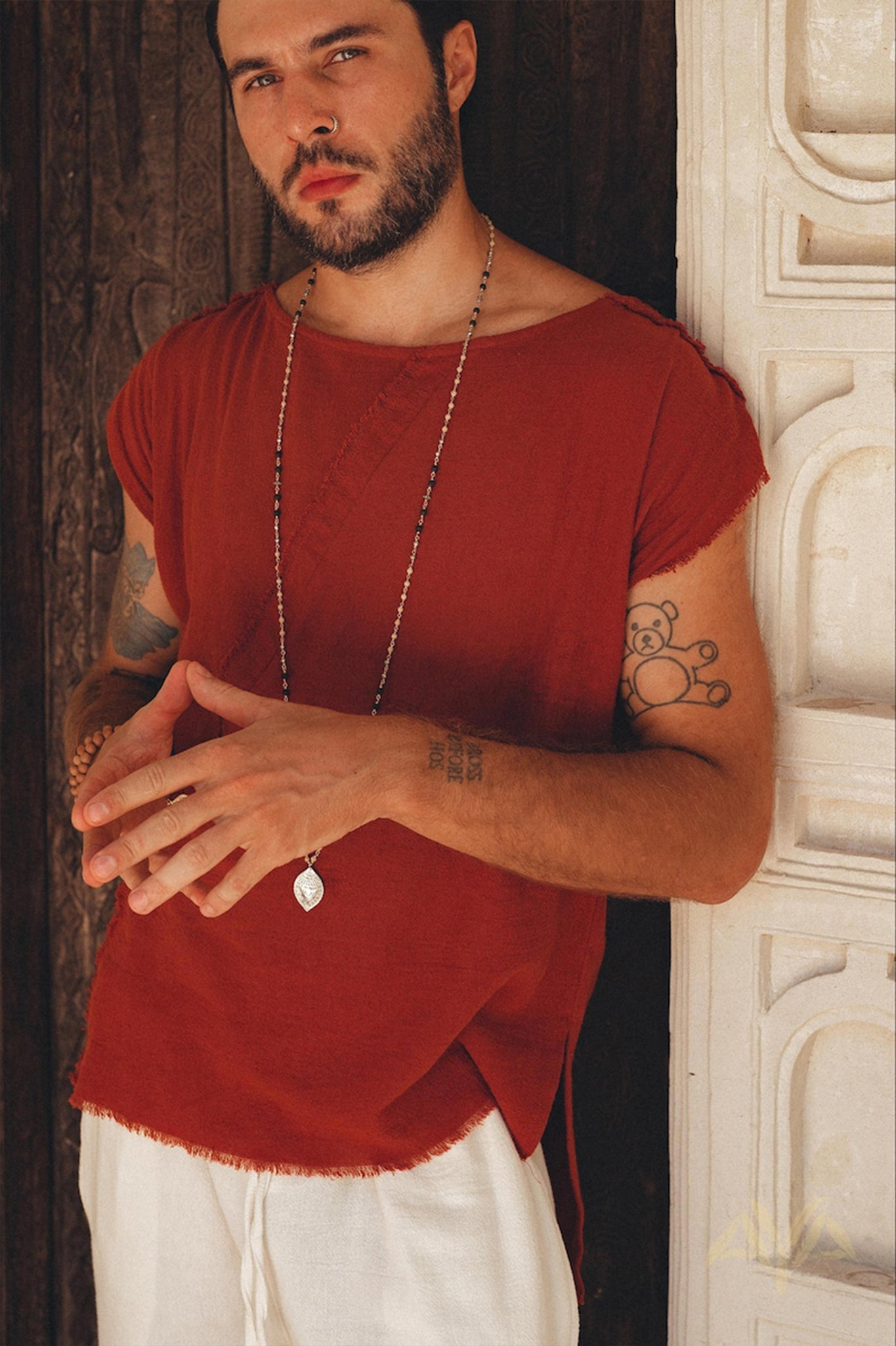 A bearded man with tattoos stands against a textured wooden wall, wearing an outfit that reflects his unique style. He dons a rust-colored shirt made of organic cotton paired with off-white handwoven cotton shorts for men from AYA Sacred Wear. To complete his look, he accessorizes with a long necklace. His hands are clasped in front of him as he gazes at the camera.