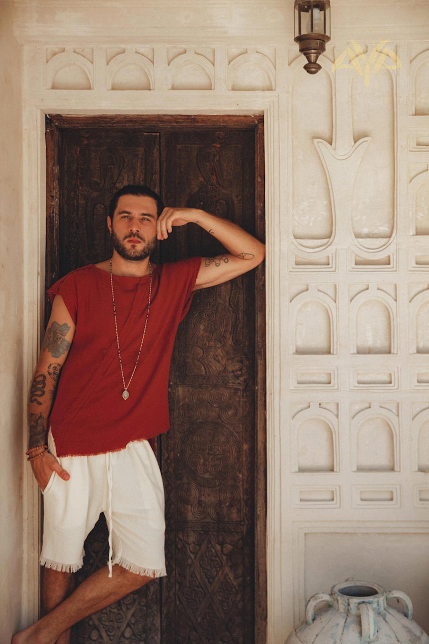 A tattooed man, dressed in a red sleeveless shirt and AYA Sacred Wear's Off-White Handwoven Cotton Shorts for Men, leans casually against a carved wooden door. He stands next to a white wall adorned with decorative patterns and has a large ceramic jar at the bottom.