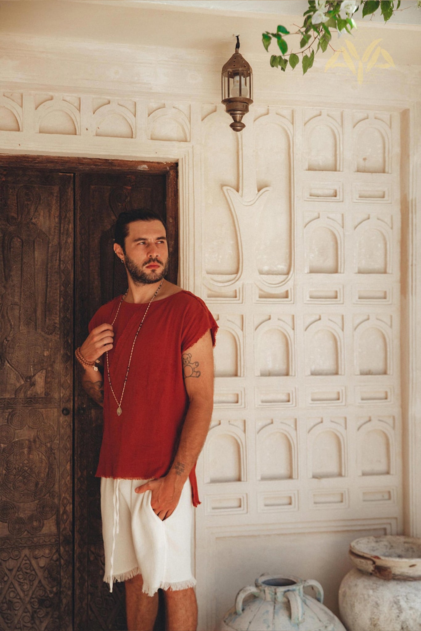 A man dressed in AYA Sacred Wear's Off-White Handwoven Cotton Shorts stands in an ornate room featuring a textured wooden door and patterned walls. A lantern hangs above him, and decorative pottery near the floor enhances the room's charm.