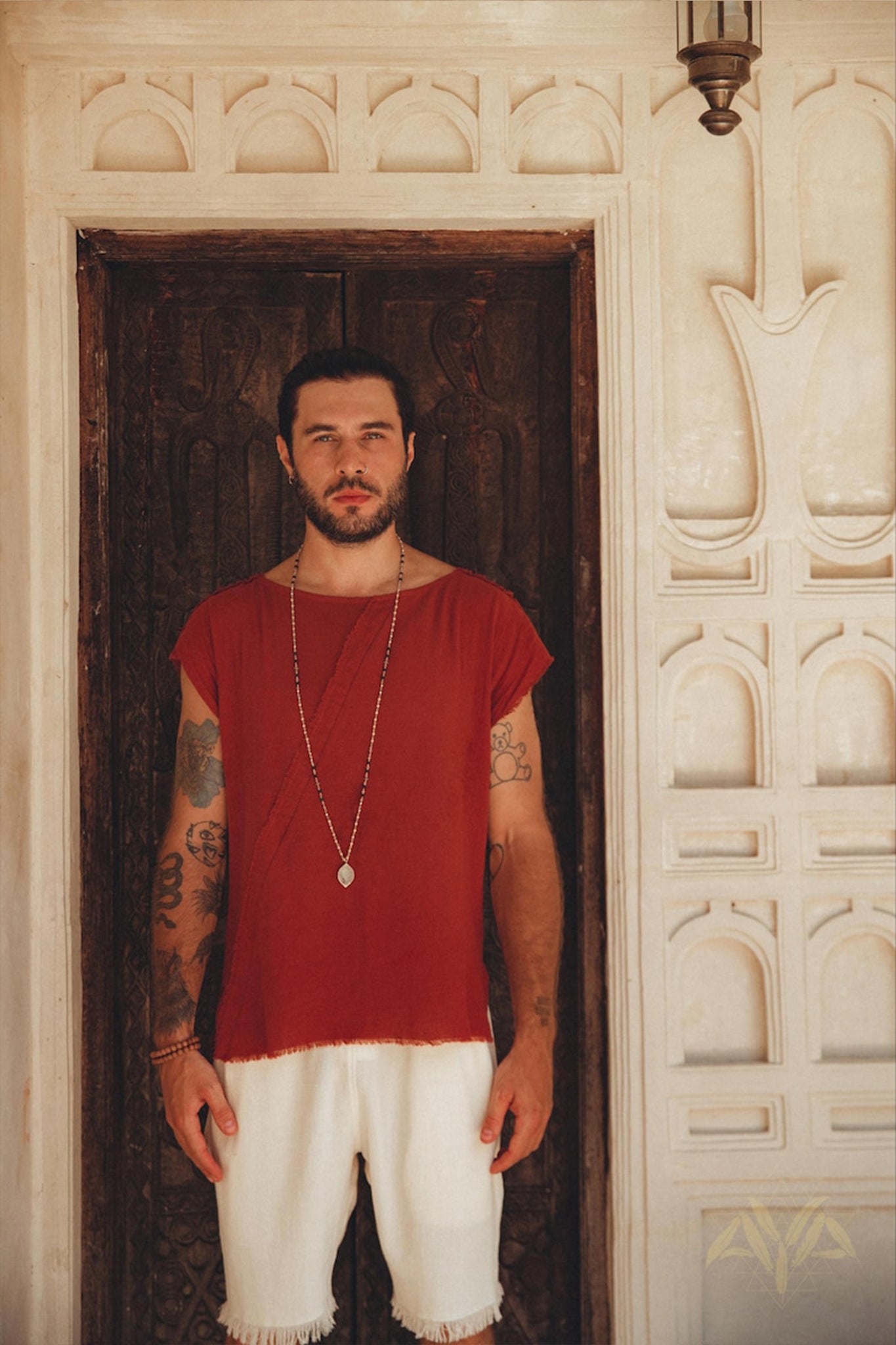 A man with a beard stands in front of an ornate wooden door and decorative wall, creating a rustic, historical ambiance. He is wearing a red shirt made from botanically dyed, organic cotton paired with AYA Sacred Wear's Off-White Handwoven Cotton Shorts for Men, and he completes the look with a long necklace.