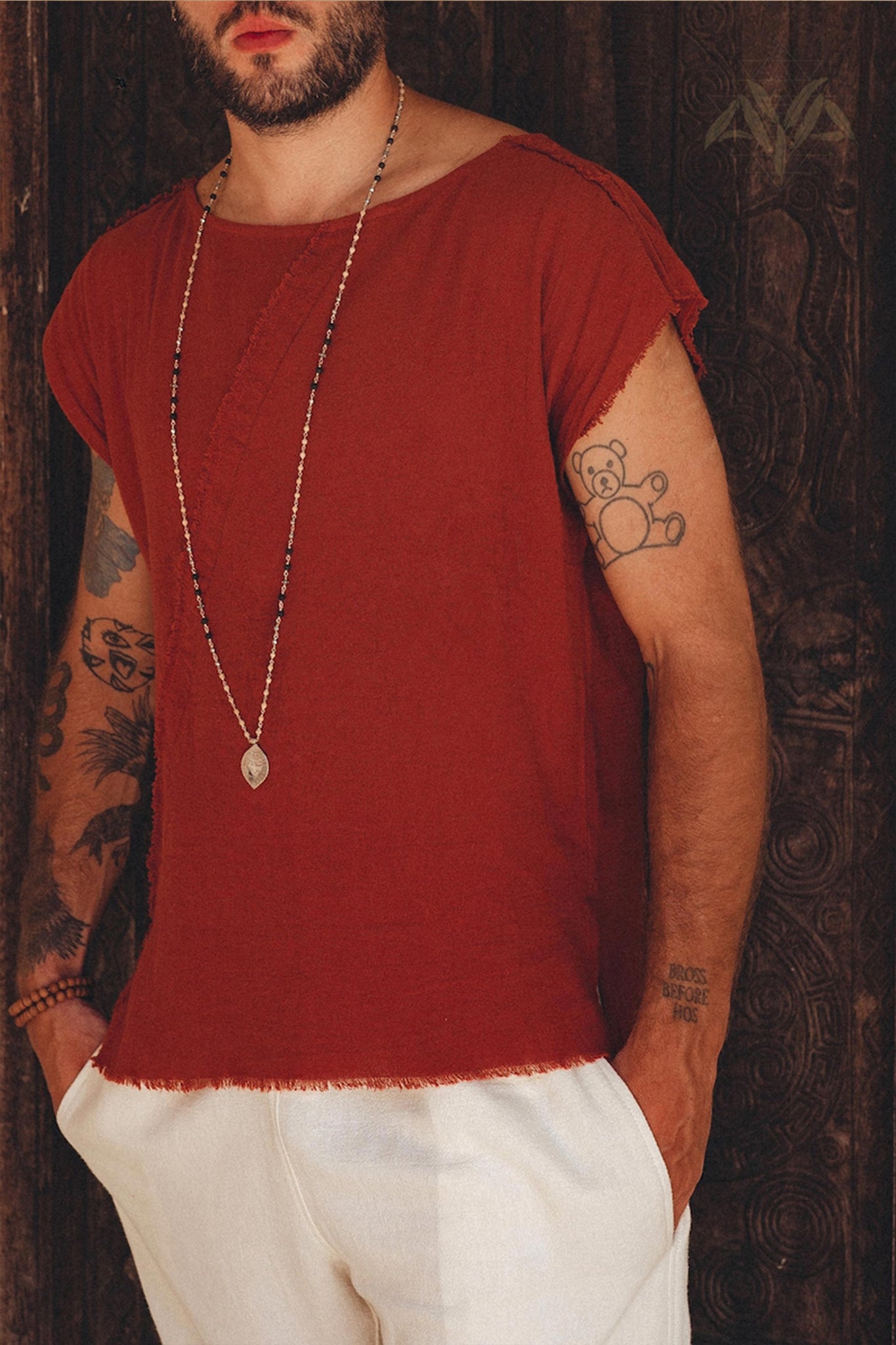 A person with tattoos on their arms is wearing a red sleeveless shirt crafted from botanically dyed organic cotton, paired elegantly with off-white handwoven cotton shorts for men by AYA Sacred Wear. Their look is accentuated by a long beaded necklace with a pendant. Dark wood with carved patterns in the background contrasts beautifully with their vibrant attire.