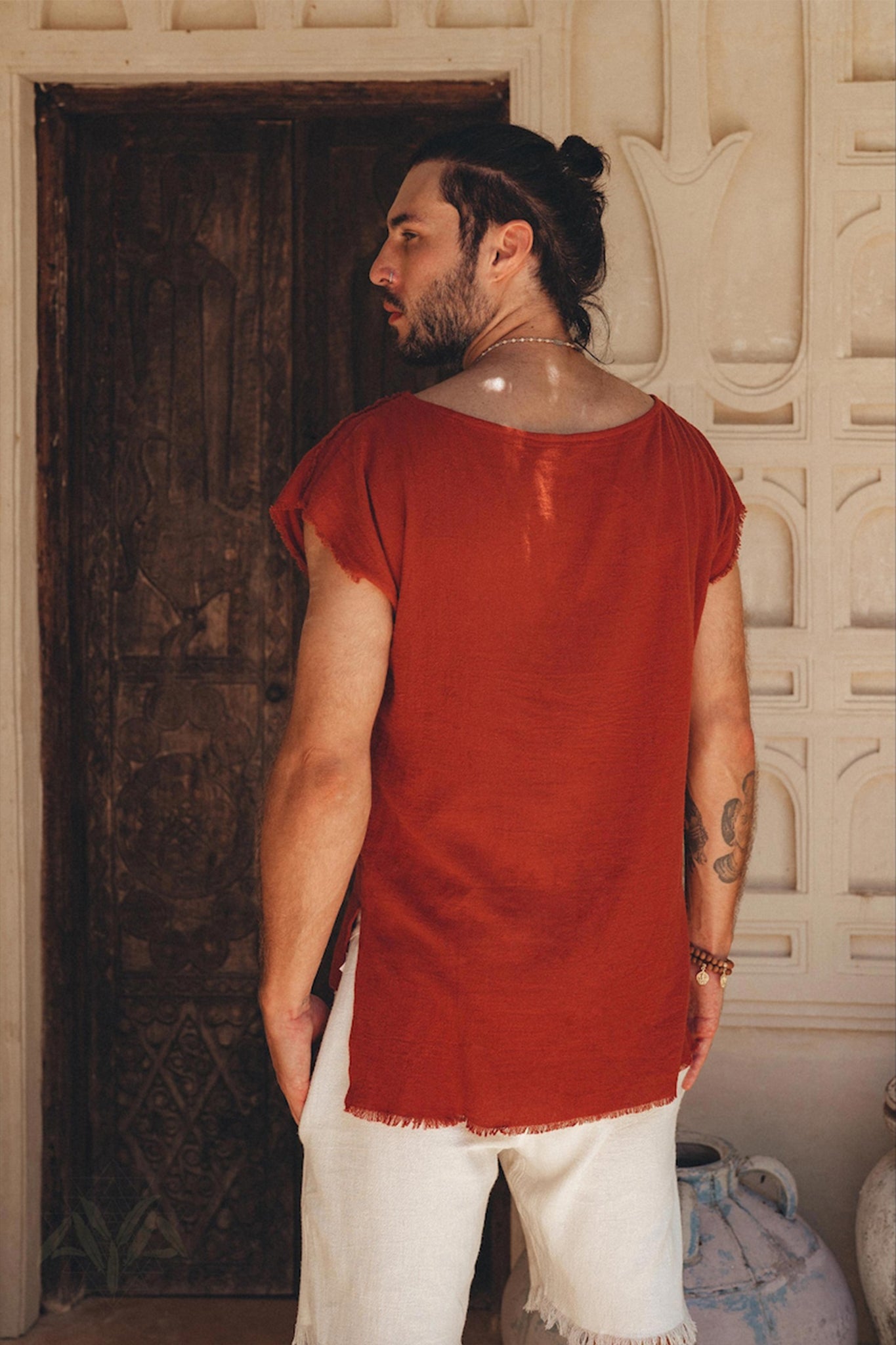 A man with a bun hairstyle, dressed in a loose-fitting red shirt and Off-White Handwoven Cotton Shorts for Men by AYA Sacred Wear, stands with his back to an intricately carved wooden door set in a cream-colored wall adorned with decorative patterns. A large pottery vase is positioned beside him.