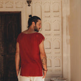 A man with long hair tied up, wearing a red sleeveless shirt and the Off-White Handwoven Cotton Shorts for Men by AYA Sacred Wear, stands with his back facing the camera. He is in front of a decorative wall with intricate patterns, surrounded by potted plants.