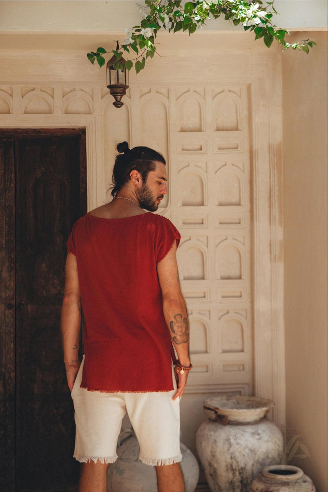 A man with long hair tied up, wearing a red sleeveless shirt and the Off-White Handwoven Cotton Shorts for Men by AYA Sacred Wear, stands with his back facing the camera. He is in front of a decorative wall with intricate patterns, surrounded by potted plants.