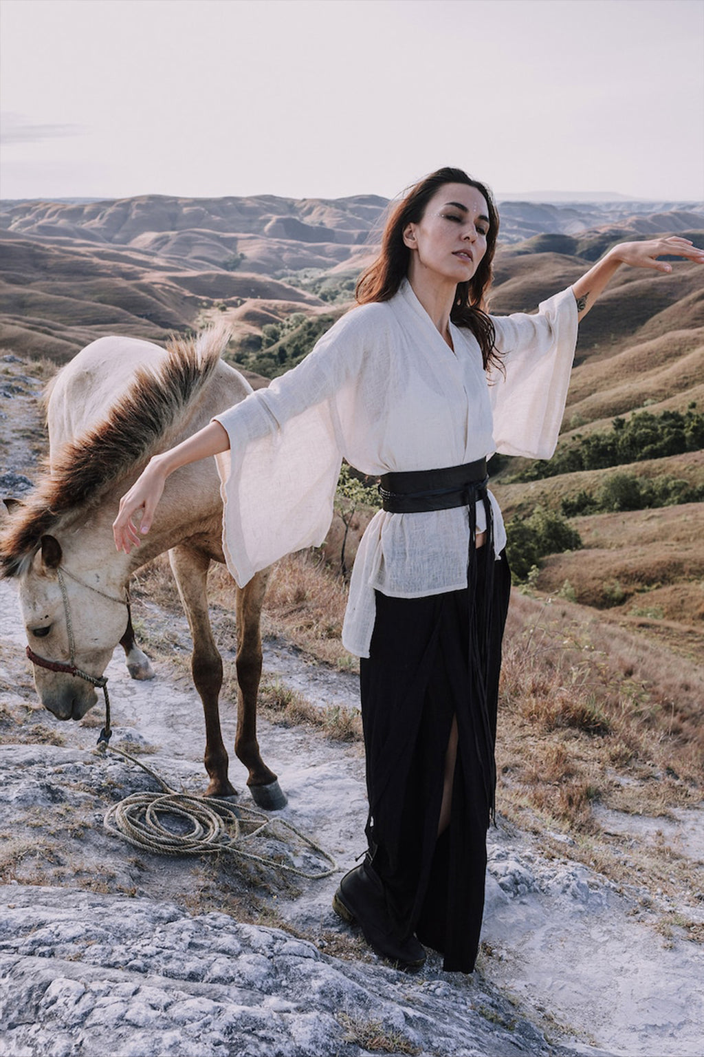 A woman in an AYA Sacred Wear Off-White Kimono Top • Tribal Boho Top and a black skirt poses with arms outstretched on a rocky hillside. A tan horse stands beside her, set against a backdrop of rolling hills beneath a clear sky.